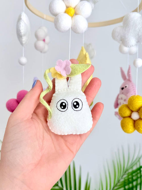 May include: A hanging mobile featuring a circular wooden frame with felt ornaments including a pink creature, a purple creature, a yellow sun with a face, white and green felt flowers, and white clouds suspended by thin white strings.
