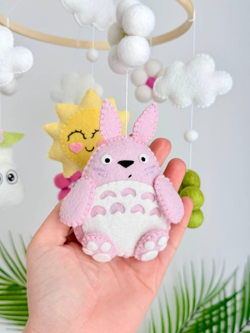 May include: A hanging mobile featuring a circular wooden frame with felt ornaments including a pink creature, a purple creature, a yellow sun with a face, white and green felt flowers, and white clouds suspended by thin white strings.