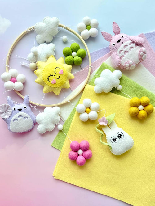 May include: A hanging mobile featuring a circular wooden frame with felt ornaments including a pink creature, a purple creature, a yellow sun with a face, white and green felt flowers, and white clouds suspended by thin white strings.