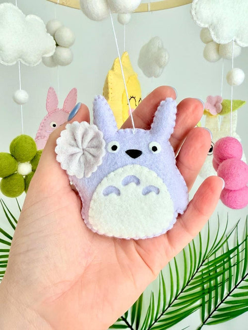 May include: A hanging mobile featuring a circular wooden frame with felt ornaments including a pink creature, a purple creature, a yellow sun with a face, white and green felt flowers, and white clouds suspended by thin white strings.