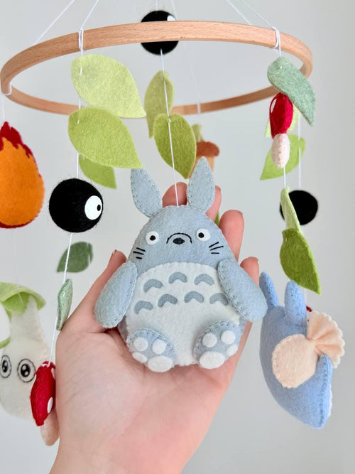 May include: A hanging mobile featuring felt figures of gray and blue creatures, a purple character, red and white spotted mushrooms, black soot sprites, and green leaves suspended from a circular wooden hoop with white strings.