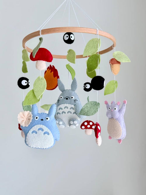 May include: A hanging mobile featuring felt figures of gray and blue creatures, a purple character, red and white spotted mushrooms, black soot sprites, and green leaves suspended from a circular wooden hoop with white strings.