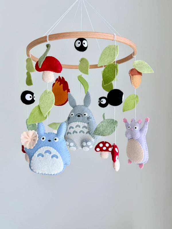 May include: A hanging mobile featuring felt figures of gray and blue creatures, a purple character, red and white spotted mushrooms, black soot sprites, and green leaves suspended from a circular wooden hoop with white strings.