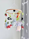 May include: A hanging mobile featuring felt figures of gray and blue creatures, a purple character, red and white spotted mushrooms, black soot sprites, and green leaves suspended from a circular wooden hoop with white strings.