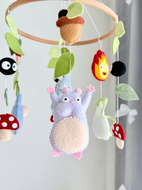 May include: A hanging mobile featuring felt figures of gray and blue creatures, a purple character, red and white spotted mushrooms, black soot sprites, and green leaves suspended from a circular wooden hoop with white strings.