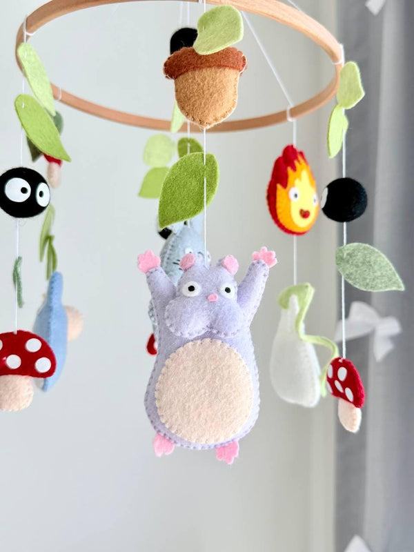 May include: A hanging mobile featuring felt figures of gray and blue creatures, a purple character, red and white spotted mushrooms, black soot sprites, and green leaves suspended from a circular wooden hoop with white strings.