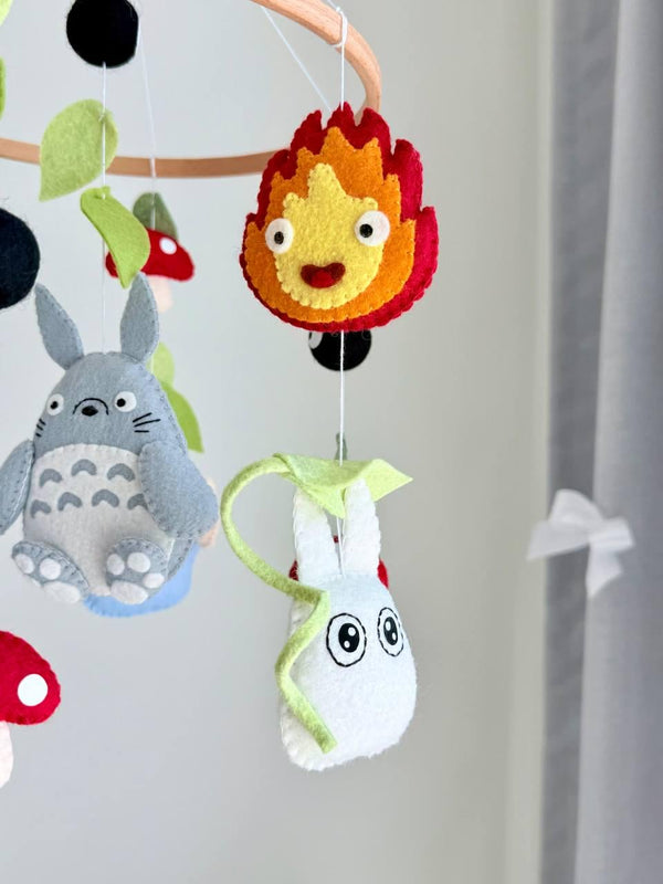 May include: A hanging mobile featuring felt figures of gray and blue creatures, a purple character, red and white spotted mushrooms, black soot sprites, and green leaves suspended from a circular wooden hoop with white strings.