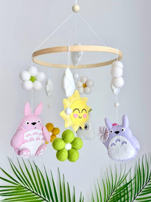 May include: A hanging mobile featuring a circular wooden frame with felt ornaments including a pink creature, a purple creature, a yellow sun with a face, white and green felt flowers, and white clouds suspended by thin white strings.