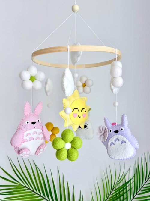 May include: A hanging mobile featuring a circular wooden frame with felt ornaments including a pink creature, a purple creature, a yellow sun with a face, white and green felt flowers, and white clouds suspended by thin white strings.